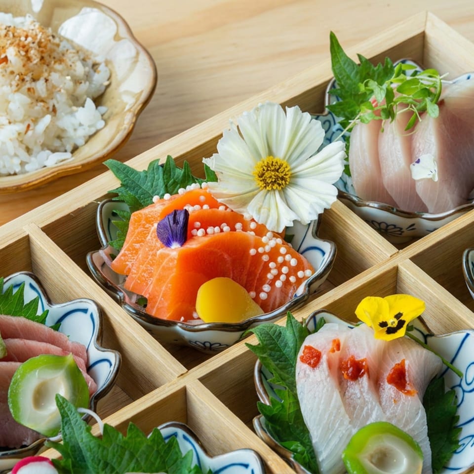 Japanese sashimi assortment arranged in individual ceramic dishes within a wooden box, accompanied by rice, shiso leaves, and edible flowers.