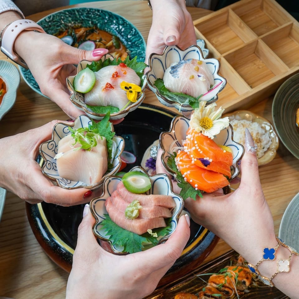 Hands present colorful sushi and sashimi dishes over a restaurant table surrounded by assorted Asian cuisine.